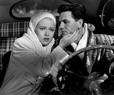 A worried woman in a headscarf and a man in a car, she gently holds his face while he drives, both in winter attire.