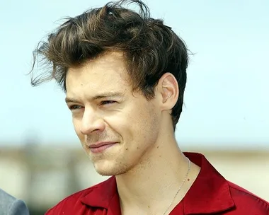 A man with wavy brown hair wearing a red shirt, smiling slightly in a bright outdoor setting.