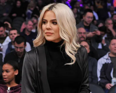 A woman with long blonde hair in a black outfit sits in a crowded stadium, looking to the side.