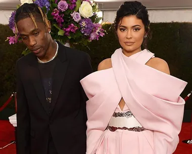 A couple poses on the red carpet; the woman wears a pink dress, and the man wears a black suit.