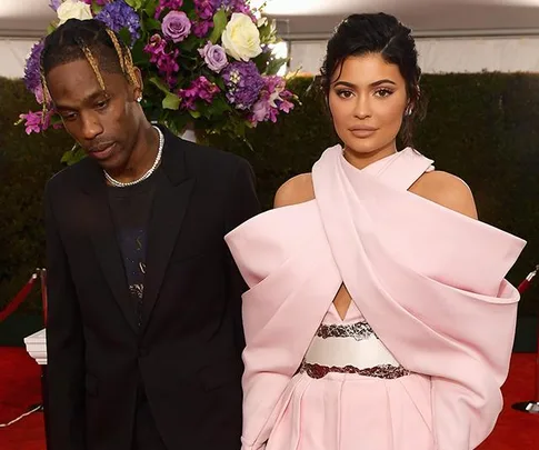 A couple poses on the red carpet; the woman wears a pink dress, and the man wears a black suit.