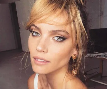 A woman with blonde hair and smoky eye makeup, wearing dangly earrings, posing indoors.