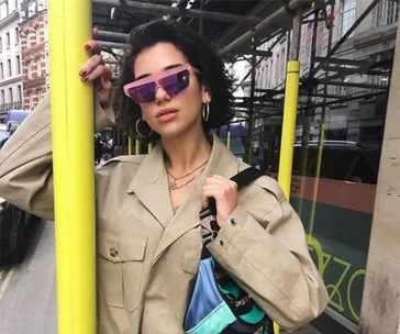 A person in a beige coat and purple sunglasses leans on a yellow pole in an urban setting.