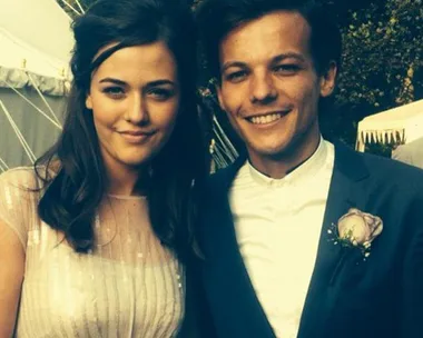 A woman in a light dress and a man in a suit with a boutonniere, smiling outdoors.