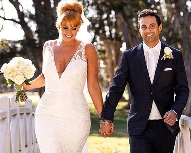 Bride in lace gown and groom in dark suit hold hands outdoors, smiling.