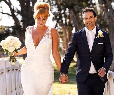 Bride in lace gown and groom in dark suit hold hands outdoors, smiling.