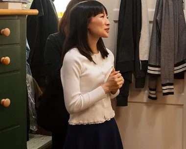 Marie Kondo in a room, smiling, wearing a white cardigan, surrounded by hanging clothes.