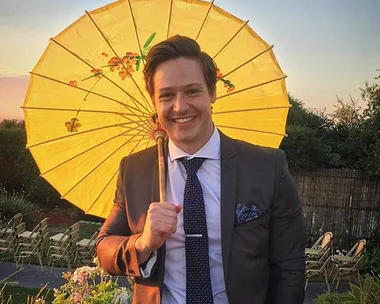 A man in a suit smiling, holding a yellow parasol with floral design outdoors during sunset.