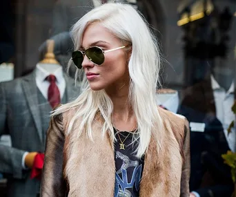 A person with long, platinum blonde hair and sunglasses, wearing a fur-trimmed jacket, stands in front of a store display.