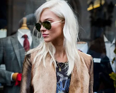 A person with long, platinum blonde hair and sunglasses, wearing a fur-trimmed jacket, stands in front of a store display.