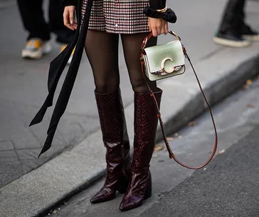 Woman wearing heeled boots, checkered skirt, holding green handbag with a "C" emblem on a city street.