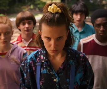 Characters from Stranger Things stand outdoors, with Eleven in focus looking determined.