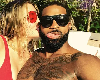 Man with sunglasses and beard, playfully sticking tongue out, with woman whispering, both in swimwear outdoors.