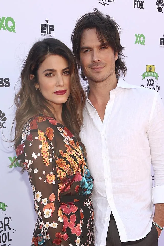 Couple posing on the red carpet; woman in floral dress, man in white shirt, logos in background.