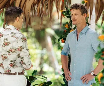 Two men standing outdoors, one in a floral shirt and the other in a light blue shirt, surrounded by tropical plants.