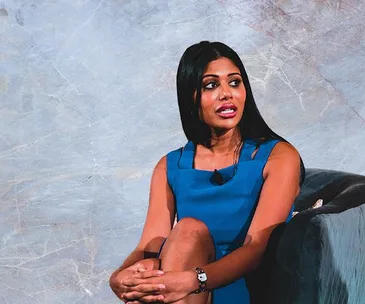 A woman in a sleeveless blue dress sits with her legs crossed, speaking in an interview or discussion setting.