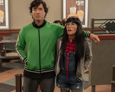 A man in a green jacket and a woman in glasses and overalls stand in a fast food restaurant.