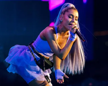 Ariana Grande performing on stage at Coachella, wearing a blue outfit and holding a microphone, with vibrant lights in the background.