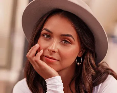 Woman with long brown hair wearing a gray hat, resting chin on hand, looking at camera with a soft expression.