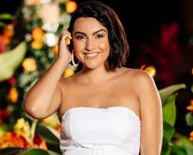 A woman in a strapless white dress smiles, standing in front of vibrant, colorful flowers.