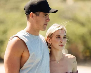 A couple stands closely together outdoors, with the man wearing a cap and sleeveless shirt and the woman in a strapless top.