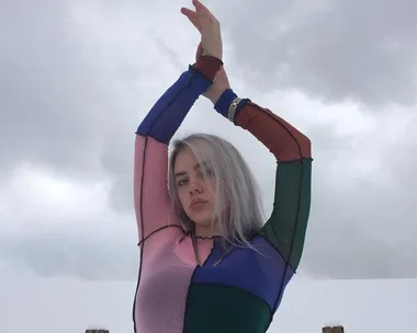 Young woman in multicolored top poses with arms raised against a cloudy sky.