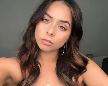 A woman with long, wavy dark hair and earrings looks directly at the camera with a neutral expression.