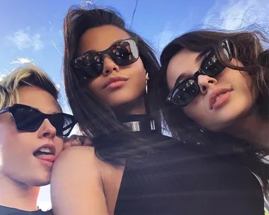 Three women wearing sunglasses pose together under a blue sky, appearing confident and stylish.