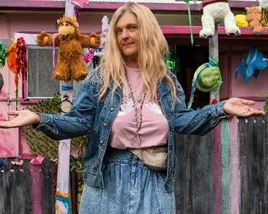 A woman in a denim outfit stands smiling, arms open, in front of a colorful yard decorated with stuffed toys and ornaments.