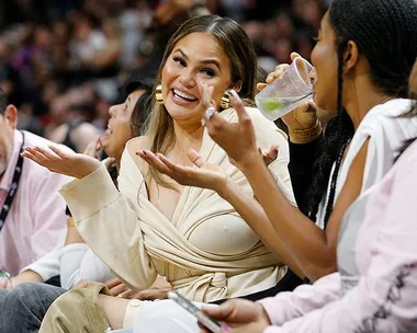 Chrissy Teigen smiling and gesturing while sitting at a sports event with friends and a crowd in the background.