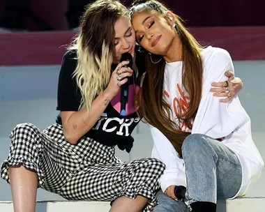 Two singers sitting closely, one singing into a mic, the other resting her head on her friend's shoulder, both appear relaxed.