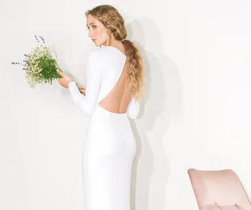 Bride in backless white gown holding flowers, standing near a pink chair, styled elegantly with long wavy hair.