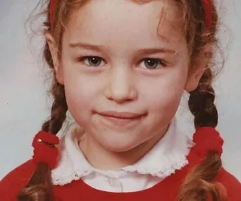 A young child with braided hair and a red headband smiles, wearing a red sweater and white collared shirt.