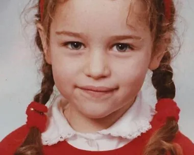 A young child with braided hair and a red headband smiles, wearing a red sweater and white collared shirt.