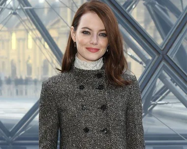 A woman with long hair in a stylish coat poses in front of a geometric glass structure, smiling.