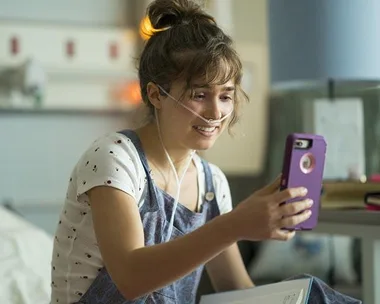 Young woman with a nasal cannula smiles at her phone, sitting in a hospital room.