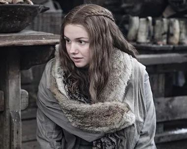 A woman with long brown hair and fur-lined cloak sits indoors, appearing thoughtful in a medieval setting.