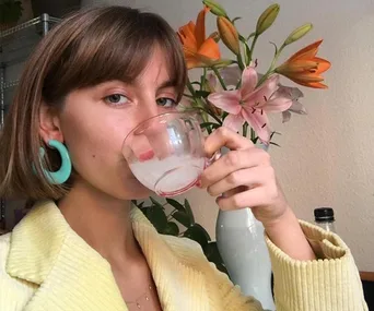 Person with short hair and turquoise earrings sips from a glass, with colorful lilies in the background.