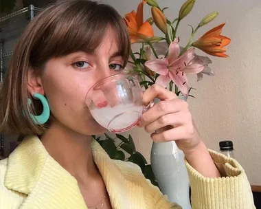 Person with short hair and turquoise earrings sips from a glass, with colorful lilies in the background.