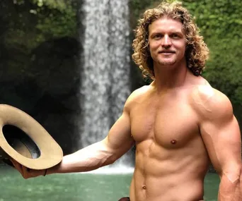 Shirtless man with curly hair holding a hat, standing in front of a waterfall in a lush, green setting.