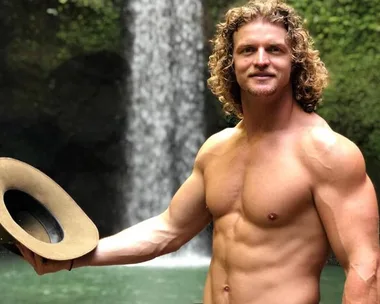 Shirtless man with curly hair holding a hat, standing in front of a waterfall in a lush, green setting.