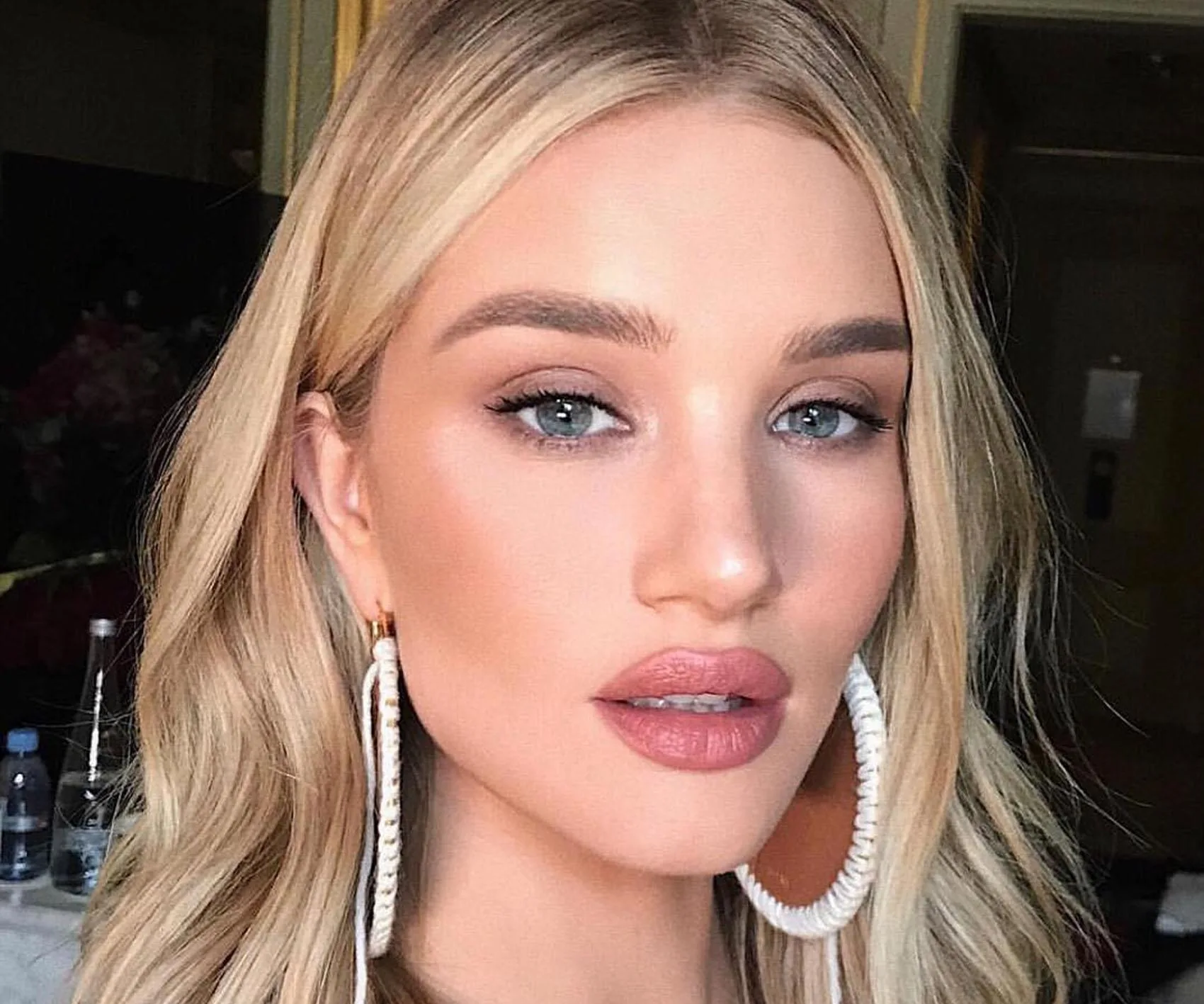 Blonde woman with flawless makeup, large hoop earrings, and glossy lips in a well-lit room.