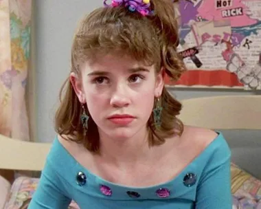 Teen girl in '80s fashion with side ponytail and blue dress, sitting in a decorated room, looking serious.