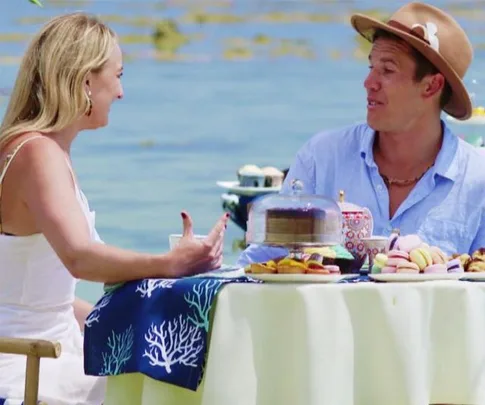 A man and a woman enjoy a seaside picnic with desserts and tea against a scenic ocean backdrop.