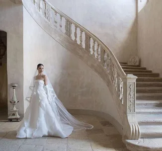 Bride in a flowing white gown stands near an elegant stone staircase, showcasing classic wedding attire.