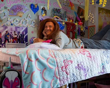 A person with long hair lies on a decorated table, surrounded by colorful posters, in a whimsical, creative room.