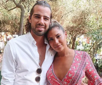 Man and woman embracing outdoors, smiling. Man in white shirt, woman in red patterned dress.