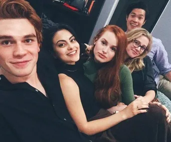 A group of five young adults, including a redhead and a man with black hair, smiling and sitting close on a couch.