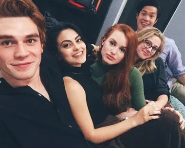 A group of five young adults, including a redhead and a man with black hair, smiling and sitting close on a couch.