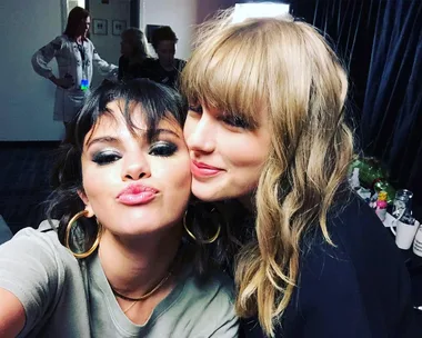 Two women happily posing for a selfie backstage, one with dark hair and the other with blonde wavy hair.